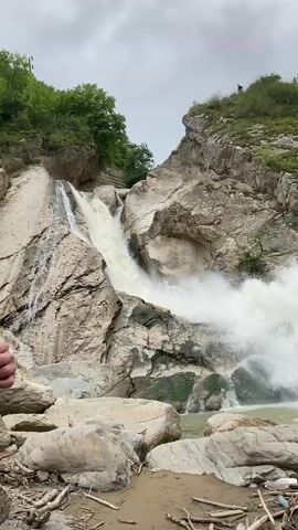The video shows a powerful waterfall cascading down a rocky cliff face. The water is turbulent and foamy as it plunges into a pool below. The surrounding environment is rugged and natural, with lush green trees covering the upper slopes of the cliffs. Several large rocks are visible in the foreground, some partially submerged in the water. There are a few people standing on a ledge at the top of the cliff, observing the waterfall. The ground near the water's edge is littered with debris, including driftwood and plastic bottles. The overall impression is one of raw, untamed natural beauty.
