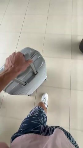 A first-person perspective video shows a person walking, pulling a grey rolling suitcase with their right hand. The person is wearing patterned shorts and white sneakers with dark green accents. The floor is covered in light-colored square tiles. The scene appears to be indoors, possibly an airport or hotel, consistent with the location context of Tambon Sa Khu, Thailand. The person's left leg is visible taking steps as they move forward. No other people or significant objects are clearly visible, except for a dark, round object in the upper right background.