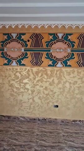 The video shows an interior space with decorative walls and ceilings. The walls are painted in two sections: the lower section has a textured gold finish, while the upper section features a colorful geometric design in shades of gold, brown, teal, and pink. A white molding with a scalloped edge runs along the top of the wall. The room has a tile floor with a marble-like pattern. There are windows with white frames and decorative iron bars. The ceiling is white with recessed lighting and decorative elements. The video appears to be a pan of the room, showcasing the interior design.