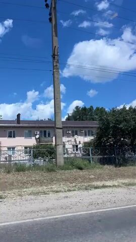 The video shows a view from a moving vehicle. The sky is blue with white, puffy clouds. There are power lines crisscrossing the upper part of the frame. In the middle ground, there are several trees, a fence, and parts of buildings, including what appears to be an apartment building. The foreground shows a dirt and gravel shoulder next to a paved road. The camera pans slightly to the right, revealing more of the buildings and trees. There are no people visible in the video. The scene appears to be in a rural or semi-urban area on a sunny day.