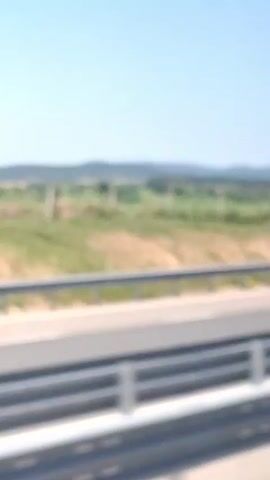 The video shows a view from a vehicle traveling along a highway in Yazdach, Bulgaria. The scene captures a bright, sunny day with a clear blue sky. The highway has multiple lanes, indicated by white lines, and is bordered by a metal guardrail.

Beyond the highway, a vast, green field stretches out, dotted with some trees and vegetation. In the distance, rolling hills and mountains are visible, covered in a mix of greenery and some sparser, drier patches, suggesting a warm climate.

A notable object in the scene is a large billboard placed on the side of the road. The billboard is red and features images of watermelons, along with text. The text appears to be in Bulgarian and reads "СУПЕР" (SUPER) and "АВТОТО" (AUTOTO). Below this, there is a logo that resembles the letter "K" inside a square, possibly indicating a brand. A silver car with a roof rack is seen driving past the billboard and the field, indicating the forward motion of the vehicle from which the video is being filmed. The overall mood is peaceful and evokes a sense of travel and open roads.