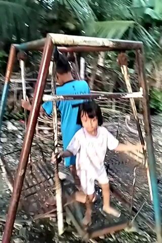 A young boy and girl are playing on a rusty swing set in a yard with tropical vegetation. The boy, wearing a blue "RONALDO" t-shirt and shorts, stands on the side, holding the frame. The girl, in white pajamas with a floral print, stands on the swing platform, looking at the camera. The swing set is old and appears to be made of metal pipes. The ground is covered with dirt and debris. In the background, there are trees, a house, and various objects scattered around the yard. The camera is handheld and shaky, creating a casual home video feel. The overall scene suggests a simple, playful moment in a rural setting.