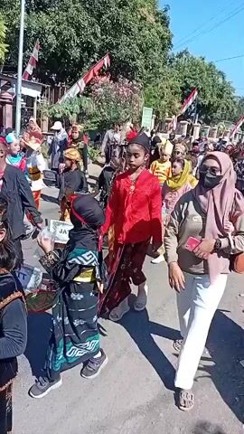 A group of children in traditional costumes walk in a parade. The children are wearing colorful outfits, including sarongs, head coverings, and decorative jackets. Some of the children are carrying signs. An adult woman wearing a hijab and a face mask walks alongside the children, holding a smartphone. Other adults and children are visible in the background. The parade appears to be taking place on a sunny day, with trees and buildings lining the street. Indonesian flags are displayed on poles in the background.