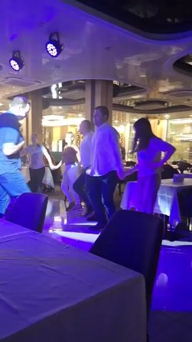 Several people are dancing in a room with tables and chairs. The room has decorative columns and a mirrored ceiling. Stage lights cycle through pink, green and blue colors. People are standing close together and dancing, facing forward. A man in a blue shirt and jeans is in the foreground. Some people are sitting at tables in the background.
