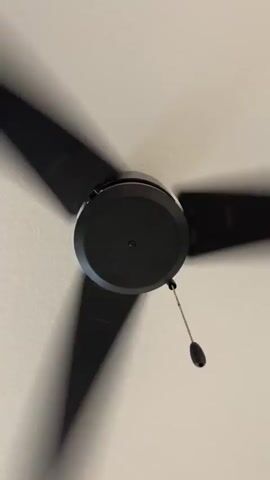The video shows a close-up, low-angle view of a black ceiling fan with three blades spinning rapidly. The fan is mounted on a white ceiling. The blades are blurred due to their speed. A thin metal chain with a black knob hangs from the center of the fan, also appearing blurred. The overall lighting is bright, suggesting it is daytime or the room is well-lit. There are no people or other objects visible in the frame. The sound of the fan is present.