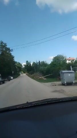 The video shows a view from inside a car driving down a paved road on a sunny day. The sky is blue with a few scattered clouds. On either side of the road, there are trees and vegetation. Some construction appears to be taking place on the right side of the road, indicated by dirt mounds and yellow caution tape. In the distance, a dark-colored car is seen approaching. The camera remains steady and focused on the road ahead. There are no people visible inside the car.