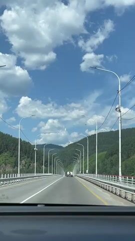 The video shows a first-person view from inside a car driving across a bridge. The bridge has a clear blue sky with white fluffy clouds above it. The bridge is lined with streetlights on both sides, spaced out evenly. The bridge has guardrails on the sides and a white line marking the lanes. There are green forested hills on either side of the bridge. Two cars are visible in the distance on the bridge. The dashboard of the car is visible at the bottom of the frame. The video is silent.