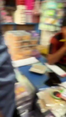 A blurry video shows the interior of a shop. In the foreground, there are shelves filled with packaged goods. In the middle ground, a person in a red shirt with black accents is seated at a counter, holding a yellow object to their mouth. Behind them, a red stool with a blue seat is visible. In the background, shelves are packed with more merchandise. The camera pans to the right, showing another person in a blue shirt standing and looking around. The shelves are filled with various items, some in plastic packaging, and boxes are stacked on the floor.