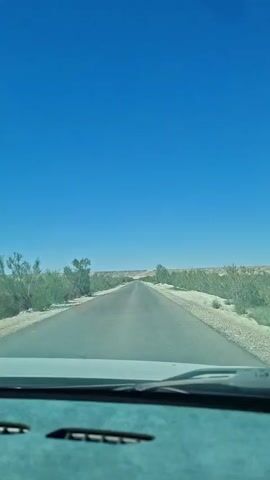 A first-person perspective video shot from inside a car shows a clear blue sky above an empty asphalt road stretching into the distance. The road is lined with dry, scrubby vegetation and sand. The car's dashboard is visible in the foreground. The video appears to be filmed on a bright, sunny day.