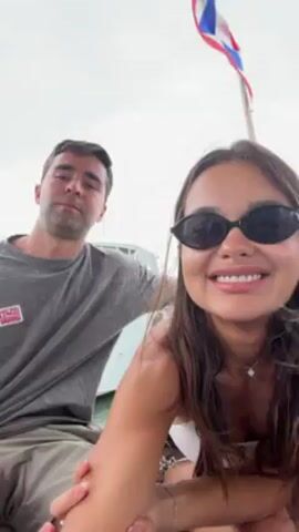 A man and a woman are taking a selfie outdoors, possibly on a boat or near a waterway. The woman, who is wearing sunglasses, is holding the camera and smiling. The man, in a grey t-shirt, is smiling as well. A Thai flag flutters in the background. The man makes a peace sign, then waves his hand over the woman's head. Both stick out their tongues and touch them together.