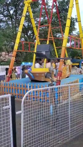 Here's a description of the video:
The video shows a rocking ship amusement park ride in operation. The ride, painted in yellow, red, and orange, is shaped like a wooden boat with a painted bow. Several children are sitting inside, wearing casual clothing. The boat swings back and forth on a blue platform surrounded by a blue and brown fence. The structure of the ride features a complex network of crossbars and ladders. A black speaker is attached to the ride's frame.

The ride is in an outdoor park setting with trees visible in the background. Other attractions and people can be seen around the ride, including a small van with flashing lights. The camera focuses on the rocking ship, capturing its motion and the children riding it. A wire mesh fence is in the foreground, partially obscuring the view.