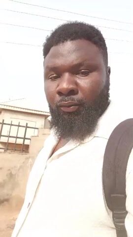 A man with a beard and short hair is walking and recording a video. He wears a white button-down shirt and a black backpack. The background includes a building, a fence, and utility lines against a bright, overcast sky.