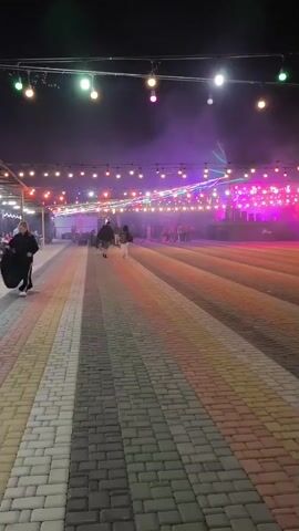 A video shows an outdoor scene at night. The ground is paved with colored bricks in stripes. String lights with colorful bulbs are hung overhead, illuminating the area with purple and pink lights. Smoke or fog drifts through the air. In the background, several people are visible, some of whom appear to be walking or standing.