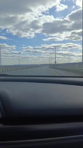 A driver’s perspective shows a highway from inside a car. The camera focuses on the road ahead, partially showing the car’s dashboard in the foreground. The highway has multiple lanes and is mostly straight, dotted with streetlights and guardrails. A truck can be seen in the distance. The sky is predominantly blue with scattered white clouds. The sun shines brightly, casting shadows on the road. The overall setting is a clear day with decent visibility.