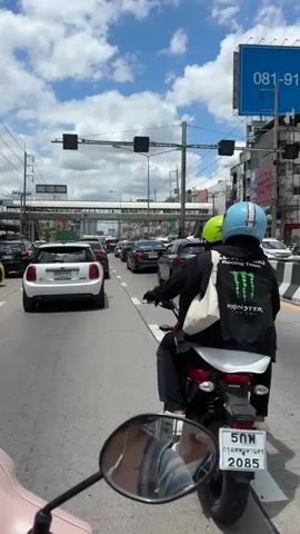 The video shows a first-person view from a motorcycle on a busy street. In the foreground, the motorcycle's mirror is visible, reflecting a person's upper body. In front of the motorcycle, there is a traffic jam with several cars and another motorcycle carrying two people. The motorcycle in front has a person wearing a black jacket with a Monster Energy logo and a light blue helmet. The other person on the motorcycle has a green helmet. Traffic lights are visible overhead, as well as a pedestrian bridge and buildings lining the street. The sky is blue with scattered white clouds. The road is paved and has lane markings.