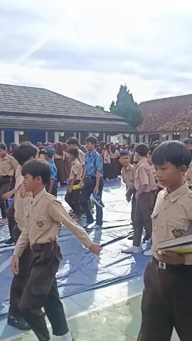 A group of students wearing scout uniforms walk around a blue tarp laid out on a paved surface. The students wear khaki shirts and brown pants, some are carrying books or papers. In the background, more students in similar uniforms are seated on the tarp. A building with a brown tiled roof is visible behind the students, along with a basketball hoop on the side of the building. The sky is overcast.