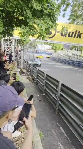 The video shows a race car speeding past spectators on a track. The car is blue and white with racing decals. The spectators are standing behind a metal barrier, and some are holding up their phones to take pictures. The track is lined with trees and a yellow banner with a black logo. The video is shot from a low angle, looking up at the race car.