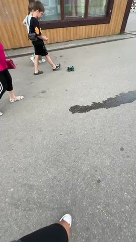 This is a short, shaky video from a low-angle perspective, likely filmed on a mobile phone. The scene is an outdoor paved area, possibly a sidewalk or parking lot, with a building in the background. The time appears to be daytime, with natural light.

The primary focus is a small, green and black remote-controlled toy car with six wheels, actively driving on the asphalt. The car is dark with bright green accents on its wheels and body.

In the background, a young boy is seen walking and operating the remote control for the toy car. He is wearing a black t-shirt with a graphic design, black shorts with white speckles, and black slides. He has a black bag slung across his chest. To his left, a girl is walking away from the camera, wearing a light green outfit and white sneakers. Further in the background, parts of a building with wooden siding and windows are visible. There is also a dark, wet patch on the asphalt.

The overall mood seems casual and playful, with the children engaged in an activity. There is no discernible text in the video.