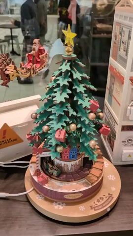 The video shows a rotating Christmas tree with a mechanical Santa Claus and reindeer flying around the tree. The tree is green and decorated with gold balls and pink presents. At the base of the tree is a small town and the words "Santa Store." The base of the tree is decorated with snowflakes and the words "Merry Christmas." The camera is static. The scene is set in a public space, with people visible in the background. To the right of the tree is a toy building. There is a white warning label to the left.