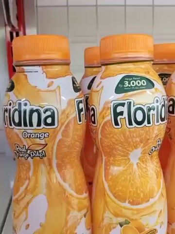 A close-up shot shows several bottles of Floridina orange juice lined up on a shelf. The bottles are clear plastic with orange caps and feature labels with the Floridina logo, images of oranges, and nutritional information. The shelf appears to be in a store or pantry, with a tiled wall visible in the background. The lighting is bright and even, suggesting an indoor setting.