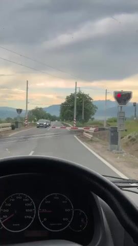 The video shows a view from inside a vehicle, looking out through the windshield at a railway crossing. The vehicle is stopped, indicated by the closed gates and a red signal light. A train passes through the crossing from left to right. The train consists of a blue and white locomotive pulling several brown freight cars. The sky is overcast with clouds. The surrounding environment appears to be rural, with trees and hills visible in the distance. The dashboard of the vehicle is partially visible in the foreground, showing a speedometer.