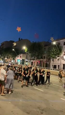 A nighttime street parade is underway. A group of people, dressed in matching black t-shirts with a green and white emblem, march in formation. Many of them are carrying what appear to be small drums or percussion instruments and some are holding white circular objects, possibly fans. The group includes men, women, and children. They are walking down a paved street lined with buildings on one side and a low barrier on the other. Streetlights illuminate the scene, and the sky is a dark blue, indicating it is dusk or early night. In the sky, several illuminated, stylized flower-shaped decorations are visible. Onlookers stand on the side of the street, watching the parade. One man in the foreground, wearing a white t-shirt and shorts, is facing away from the camera, looking towards the parade. The overall atmosphere appears festive.