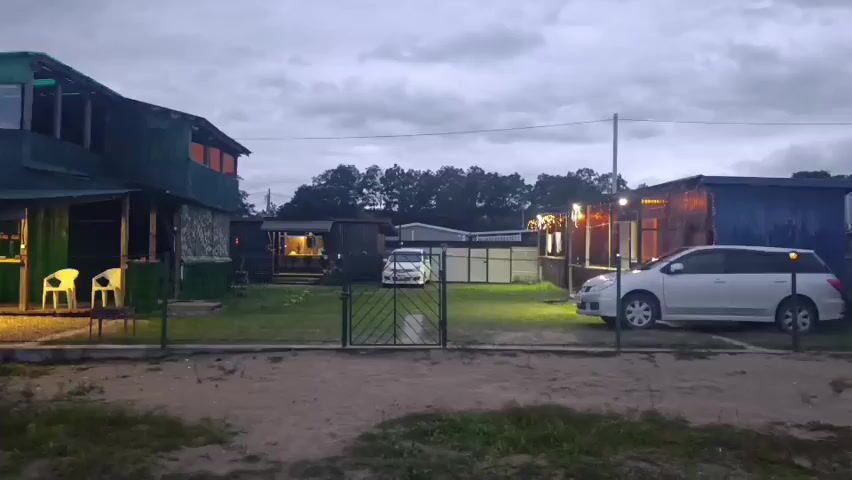 The video shows an outdoor scene at dusk. There are two buildings visible. The building on the left is a two-story structure with a green metal roof and what appears to be an open-air section on the first floor. Two white chairs are positioned outside this building. The building on the right is a single-story structure with a blue metal exterior. Two white cars are parked in the yard. The ground is covered with grass and some sand. A metal gate is visible in the foreground. The sky is overcast.