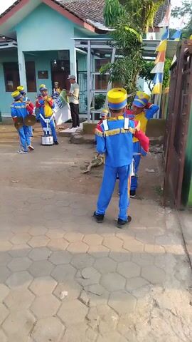 A group of young people in matching blue and yellow marching band uniforms are gathered outdoors. Some are holding drums and cymbals, while others are holding flags. One individual in the foreground is holding a baton. They appear to be preparing for a performance. The background shows a building with a red roof and a flag pole with a red and white flag. There are also trees and motorcycles visible. The ground is paved with hexagonal stones.