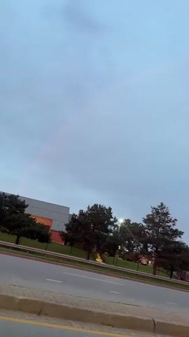 This is a vertical, outdoor, medium shot video taken from a car moving along a road. The sky is overcast with a hint of a rainbow visible on the left side of the frame. On the left side of the road, there is a building with an orange accent and some trees. Further down the road, there are several cars driving in traffic. The cars are mostly dark in color, and their brake lights are visible. A "ONE WAY" sign is visible on a pole to the right of the road. The video has a slight motion blur due to the movement of the car.