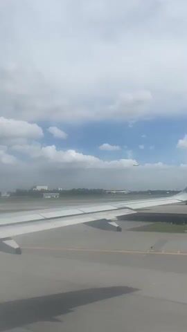 The video shows a view from inside an airplane, looking out of a window. The plane's wing is visible in the foreground. Outside, the sky is partly cloudy with patches of blue sky. Another airplane is descending in the distance, approaching the runway. The ground visible beneath the plane and in the distance includes what appears to be an airport runway and buildings, interspersed with greenery.