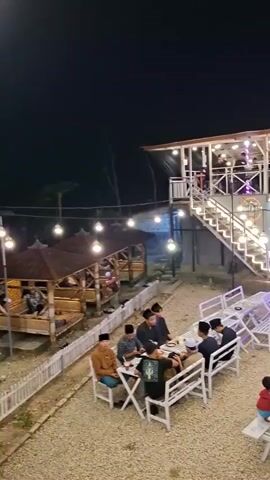 The video pans across an outdoor seating area at night, illuminated by strings of hanging light bulbs and lights from a two-story building. Several groups of people are seated at tables, some at picnic tables with benches and others at smaller tables with chairs. Some individuals are wearing traditional Islamic clothing. The ground is covered in gravel. In the background, motorcycles are parked near the building. The building has an open-air second floor with railings and more lights. The overall scene appears to be a casual dining or gathering spot.