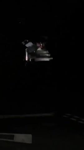The video shows a dark road at night. There are faint lights visible in the distance, possibly from a vehicle or buildings. The camera is moving, suggesting it is attached to a vehicle. There are some white markings on the road in the foreground, indicating a lane divider or crosswalk. The overall scene is dark and somewhat ominous.