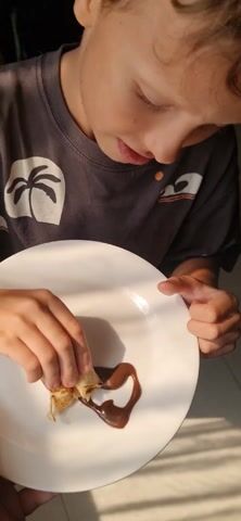 A young boy is shown eating a piece of pancake with chocolate spread. He is wearing a dark grey t-shirt with a palm tree and wave graphic. He dips a piece of the pancake into the chocolate, which is arranged in a heart shape on a white plate, and eats it. The boy is fair-skinned and has short, light-colored hair. The setting appears to be indoors, possibly near a window, given the soft light.