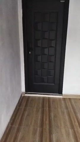 The video pans from left to right, showing a dark-colored door with a geometric pattern. The door is closed and appears to be made of metal. To the left of the door is a white wall and to the right is another white wall with a dark rectangular opening, possibly a window or a ventilation shaft. The floor is covered with wooden planks.