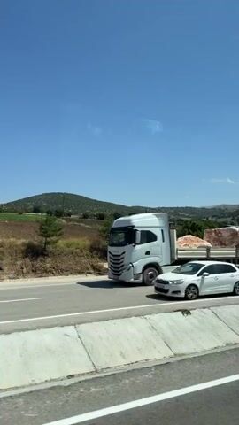 A white pickup truck drives down a highway. The truck is the only vehicle visible on the road. The truck is traveling in the right lane. The road is paved and has a white line marking the lane. The highway is surrounded by green hills and trees. The sky is clear and blue. The video is taken from a moving vehicle, possibly another car or a bus, as evidenced by the reflection in the window.