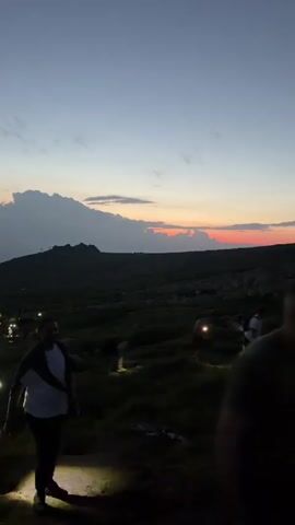 The video shows a group of people hiking on a hillside at dusk. The sky is a deep blue with streaks of orange and pink from the setting sun. Below, a city twinkles with lights. The hikers are using headlamps to illuminate their path as they ascend the rocky terrain. The mood is one of adventure and perseverance as they navigate the darkening landscape.