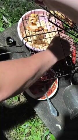 A close-up overhead shot shows a person's hands preparing food outdoors. In the foreground, a metal grill basket is positioned over a wooden surface. The basket contains grilled sausages, which appear to be sliced and are resting in a red sauce on a plate with a fork. To the left, a pink bowl holds grilled chicken pieces and a pile of shredded food, possibly noodles or cabbage. The background consists of green grass and foliage, suggesting an outdoor setting. The lighting is dim, indicating it might be evening or nighttime. The person's hands are actively manipulating the grill basket and the food.