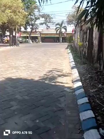 The video shows a paved area with brick paving stones. To the right, there is a concrete wall with some greenery growing along it. On the left side of the paved area, there are trees and parked cars. In the background, there is a building with a sign that appears to say "Pangsit Mie Ayam". There are several people on motorcycles moving in the distance. The sky is clear and the sun is shining, casting shadows on the paved area. The camera is held steady and appears to be filming from a stationary position.