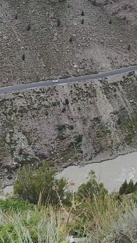 A white vehicle travels on a paved road that winds along the side of a steep, rocky, and sparsely vegetated mountain. Below the road, a wide, muddy river flows through a valley. The foreground is dominated by lush green grass and some thorny plants. The overall scene depicts a remote mountain landscape with a road and river.