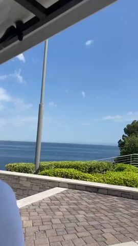 This is a video taken from a balcony or patio overlooking a bright, sunny body of water, likely the Adriatic Sea given the location context. The scene is a scenic coastal view. In the foreground, there are architectural elements of a building, including a textured wall, stone steps, and a white railing. Lush green bushes are planted along the terrace, providing a vibrant contrast to the stone.

The middle ground is dominated by the calm, blue sea, which stretches out to the horizon under a clear, bright blue sky dotted with a few wispy white clouds. A tall, grey pole, possibly a flagpole or a light post, stands prominently in the water or on the shoreline in the early part of the video. Further out in the sea, there are faint outlines of boats or distant landmasses. The shoreline is rocky in places, with areas that appear to be designated for relaxation, possibly with beach umbrellas and chairs visible. A large umbrella, likely a patio umbrella, is visible in the frame in the latter half. A mature tree with dark green foliage is seen on the right side, its branches reaching out towards the sea.

The weather appears to be warm and pleasant, indicative of a sunny day, likely late morning or early afternoon given the brightness and angle of the sun. The overall mood is one of tranquility and relaxation, suggesting a vacation or a peaceful day at a resort. There is no visible text within the video. The camera is panning slowly across the scene, offering a panoramic view of the coastal landscape.