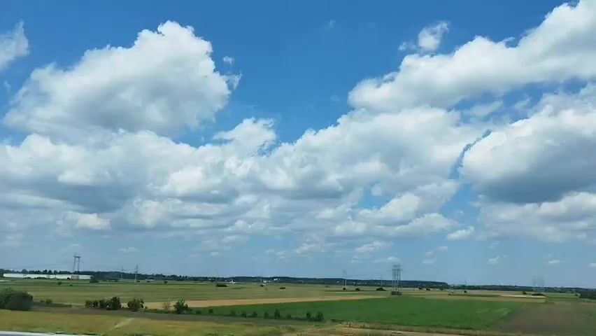 The video shows a time lapse of the sky above a rural landscape. The sky is a vibrant blue with large, fluffy white clouds drifting by. Below the sky is a vast expanse of green and yellow fields, interspersed with scattered trees and some distant buildings. There are also several tall, skeletal structures that appear to be power lines or transmission towers dotted across the horizon. The lighting suggests it is daytime, likely a sunny day with a few clouds. The scene conveys a sense of open space and nature.