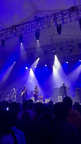 A band performs on stage for a crowd. The band consists of five members: a drummer on the left, three guitarists across the front, and another guitarist on the right. The stage is lit with bright white and blue spotlights. A grid of lighting fixtures hangs above the stage. The audience is dark and mostly obscured, but faces can be seen here and there. Some people hold up phones. A person in the foreground wears a yellow shirt with the word “PIXIES” on it. The band performs under a large tent.