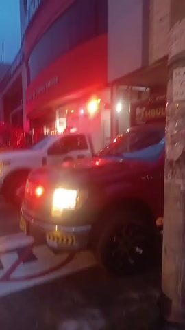 The video shows multiple emergency vehicles, including fire trucks and an ambulance, parked in front of a fire station. The station has a red and white sign that reads "BOMBEROS RUMINAHUI CENTRO DE CAPACITACION." The vehicles have their emergency lights activated, flashing red and white. The sky is a dark blue, suggesting it is either dawn or dusk. There are lane markings on the ground. The scene is likely depicting emergency responders preparing to leave the station.