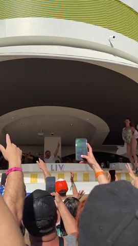 A crowd is holding up phones and recording an event. Two DJs are visible on a raised platform with their arms raised in the air. One DJ wears sunglasses and a black t-shirt. The other DJ wears a white t-shirt. A woman in a green outfit and a light-colored wrap stands off to the side. The backdrop of the area is a yellow and white striped pattern. The ceiling above the DJs has recessed lighting and a video projector.
