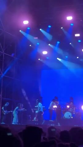 A band performs on a brightly lit stage in front of a crowd. The stage is illuminated with bright white and blue spotlights, and a large screen displays a red geometric horse head against a blue background. The band consists of a keyboardist, guitarist, drummer, and what appear to be two vocalists or performers, all silhouetted against the bright lights. The crowd is only visible in the foreground as dark silhouettes. The stage structure appears to be a metal scaffolding.