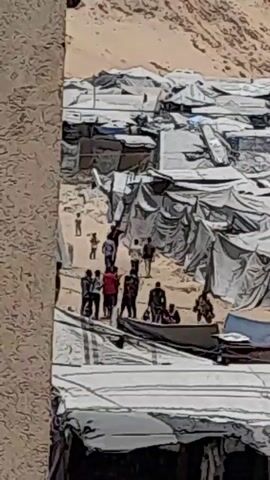 A group of people are gathered in a semi-enclosed area. Some individuals are standing and interacting. There are various structures made of tarps and other materials, suggesting a makeshift camp or settlement. A boat is visible in the foreground, with some people in it. The ground appears to be sandy or dusty.