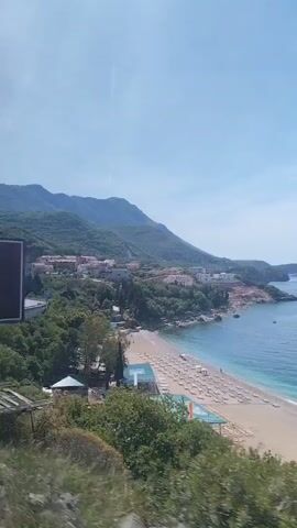 The video shows a coastal landscape from a moving vehicle. In the foreground, there's a billboard advertising "Tehno Max" with a map. Beyond the billboard, the view opens up to reveal a sandy beach dotted with beach umbrellas and a few people. The turquoise water of the sea stretches out, meeting the horizon under a clear blue sky with a few wispy clouds. In the background, there are mountains covered with greenery and a few buildings nestled along the coastline. As the vehicle moves, the view shifts, with the lush greenery of trees taking up more of the frame.