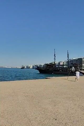 The video shows a body of water and a sandy area. On the water, there is a dark-colored boat with masts, resembling a pirate ship. The boat is docked or anchored near a city skyline visible in the distance. The sky is clear and blue. A person is visible on the sandy area near the water, standing. The camera is tilted at an angle.