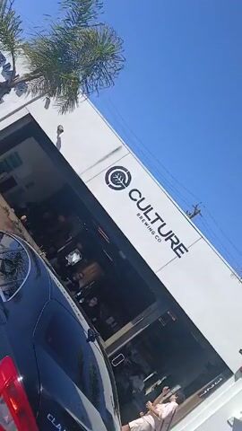 The video depicts a street scene on a sunny day. Parked cars line the street in front of a building identified as "Culture Brewing Co." Palm trees dot the landscape. A few people are seen walking near the building. The video is taken from the perspective of someone inside a vehicle.