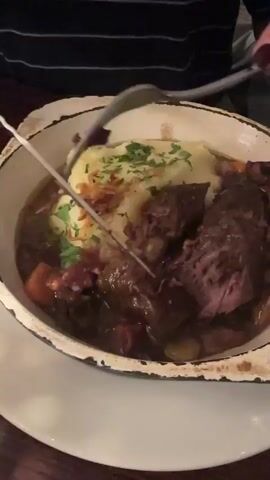 A person is cutting a dish of beef stew and mashed potatoes. The beef stew has a dark brown gravy, and visible chunks of meat and vegetables. The mashed potatoes are topped with parsley. The dish is in an enamel coated pan, sitting on a white plate on a wooden table. The person is wearing a striped shirt. They are using a knife and fork to cut a piece of beef stew.
